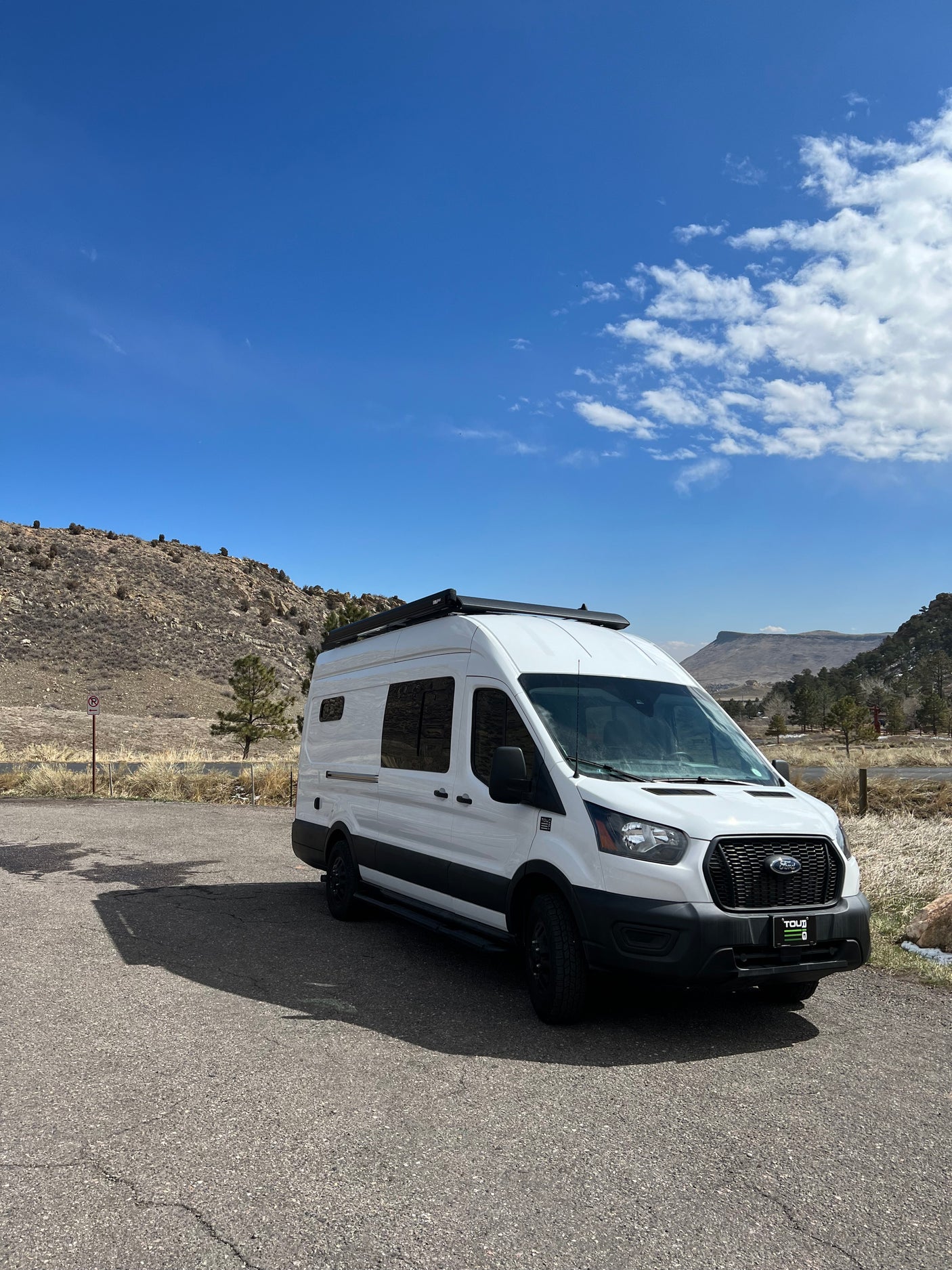 White Ford Transit campervan RV rental with roof awning, parked on asphalt in a scenic mountainous landscape under a blue sky.