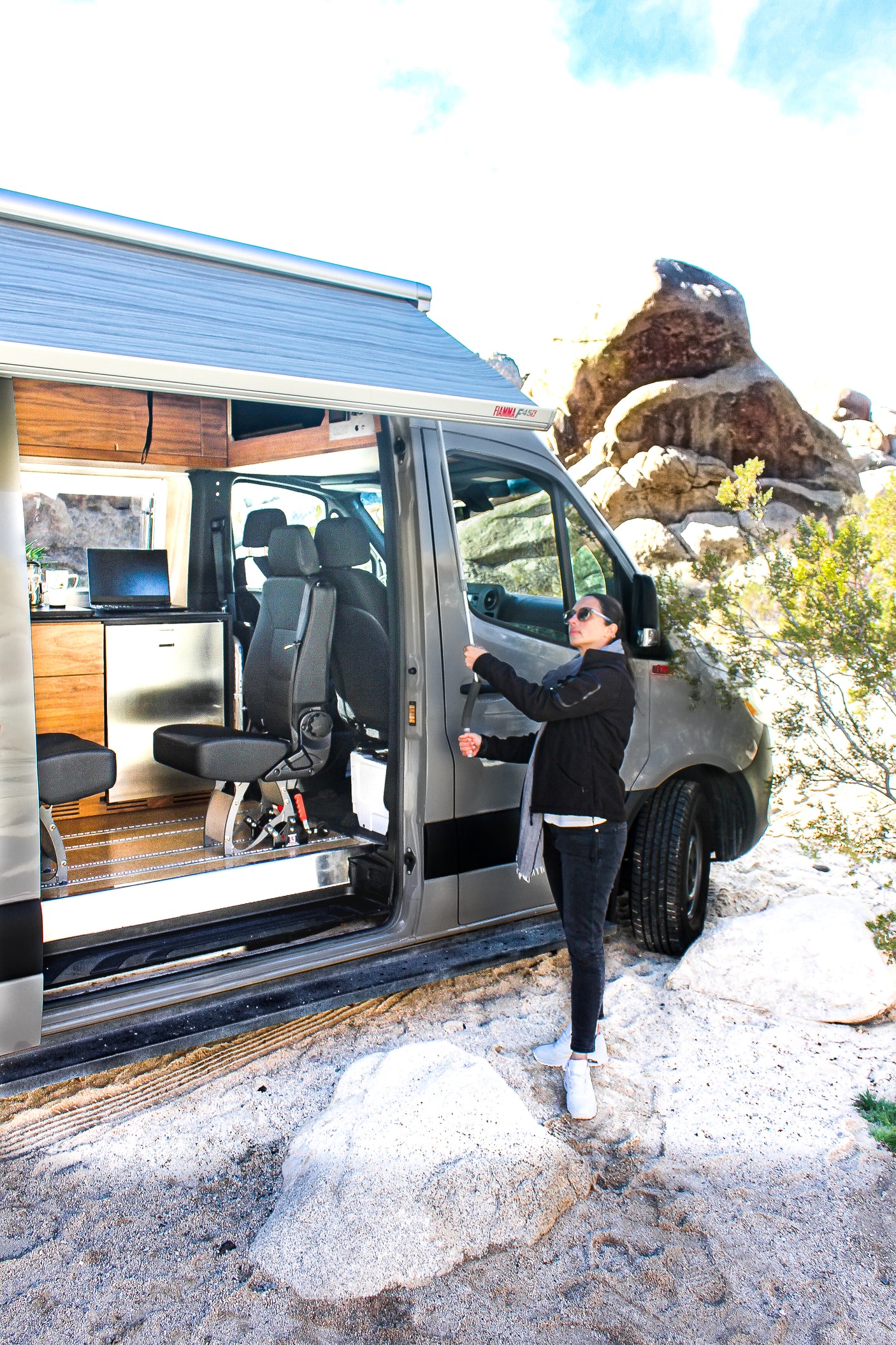 Experience van life in an RV rental. Woman by an open camper van with an awning, kitchen, and seats in a desert landscape.