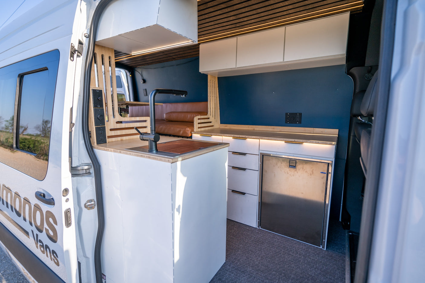 Step into this spacious RV rental camper van, featuring a modern kitchen with sink, fridge, and comfy seating.