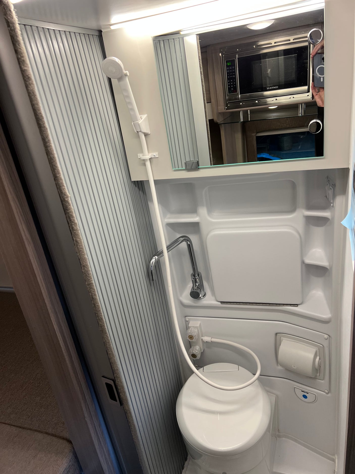 Inside an RV rental: a compact wet bath with toilet, shower, sink, and a mirrored cabinet reflecting the kitchen microwave.