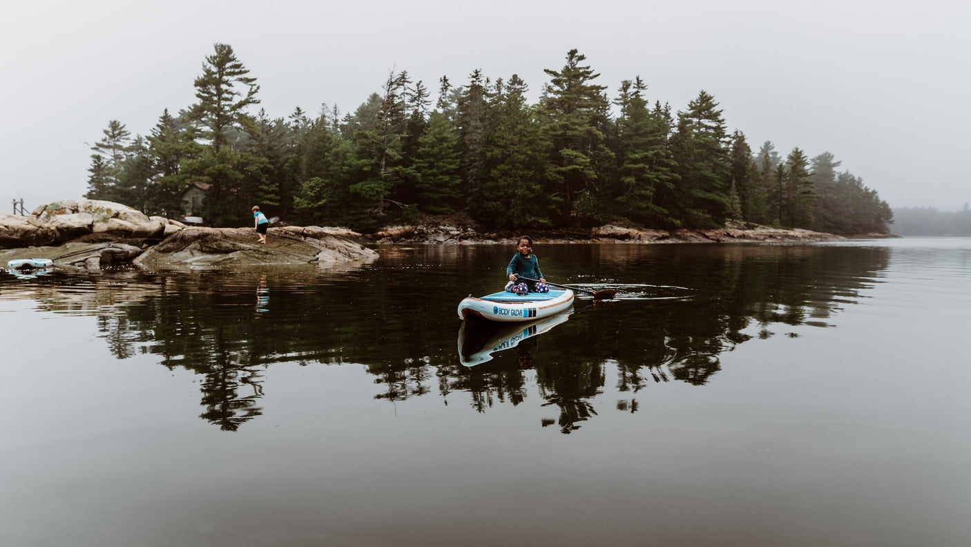 Misty lake tranquility: a child paddles on a SUP, another explores rocky shore near a forest. Ideal for an RV rental adventure!