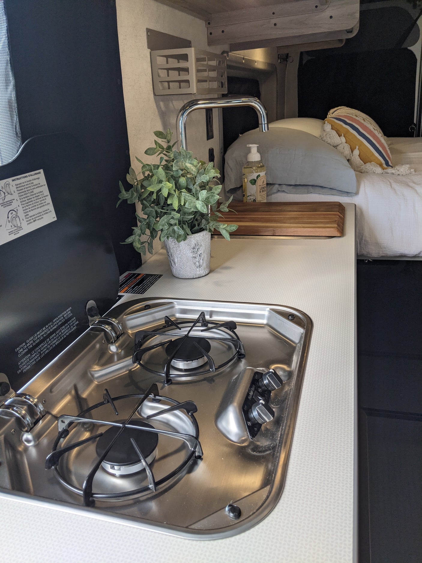 Modern RV rental: a compact kitchen setup with a stainless steel two-burner stove, sink, and a comfortable bed.