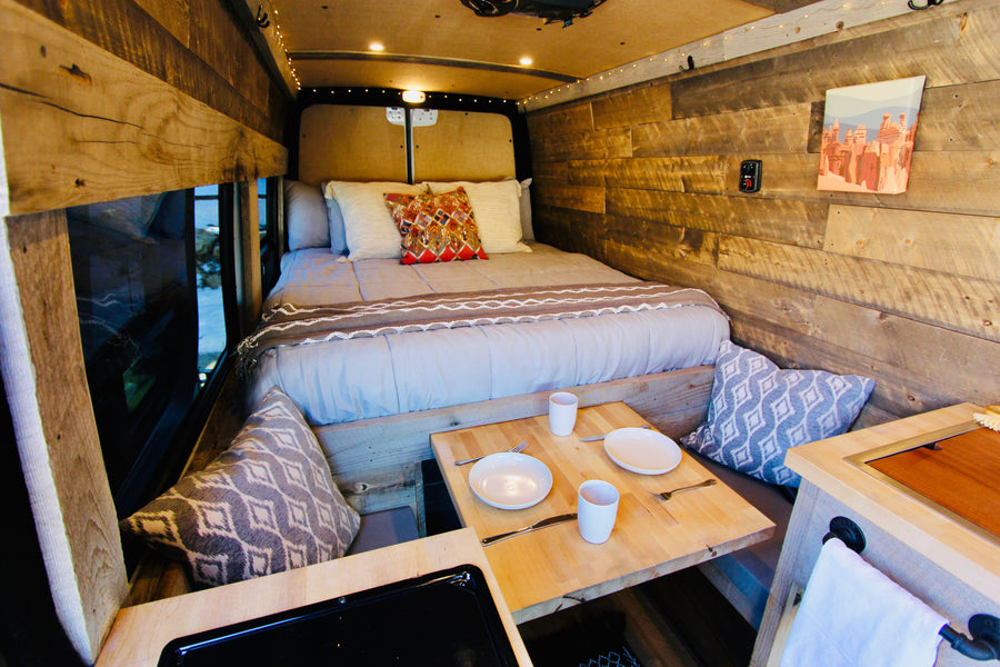 Cozy rv rental interior: a comfortable bed, rustic wooden walls, and a small dining table with place settings. Ideal camper comfort.