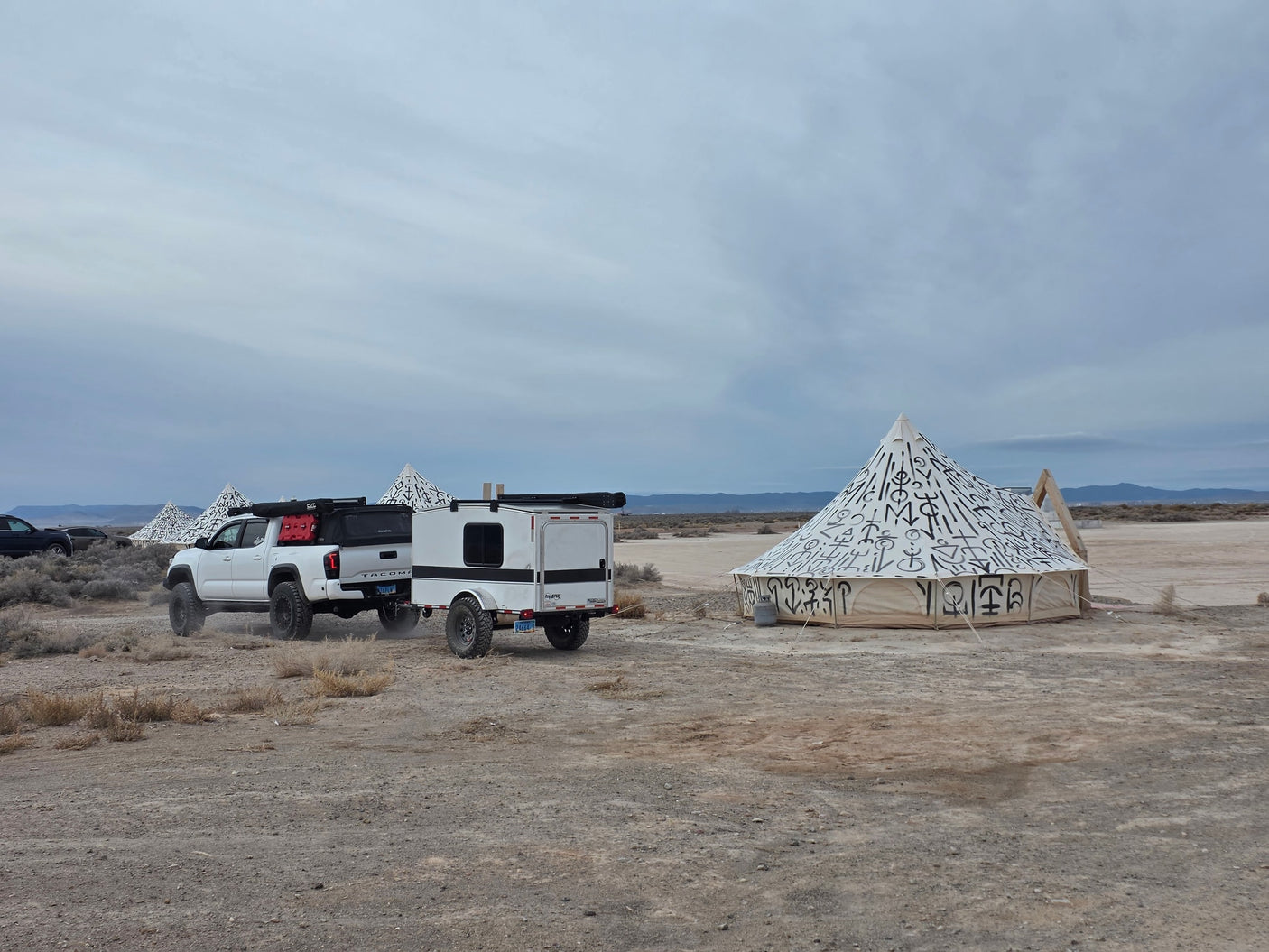 Adventure-ready RV rental: White Tacoma tows an overland trailer past art-covered tents in a dusty desert landscape.|Overland RV rental setup with white pickup truck