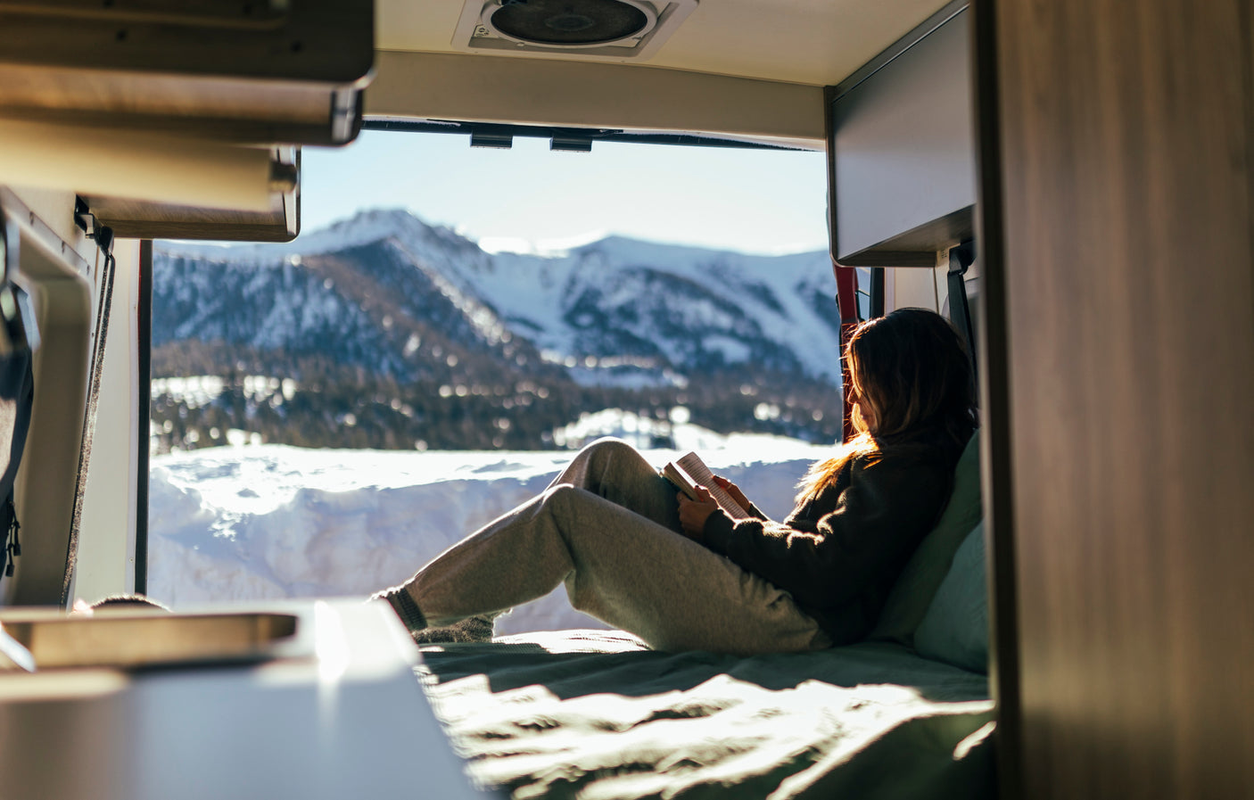 A woman relaxes in her rv rental, reading a book with a snowy mountain view from her cozy, sunlit interior. Winter escape.