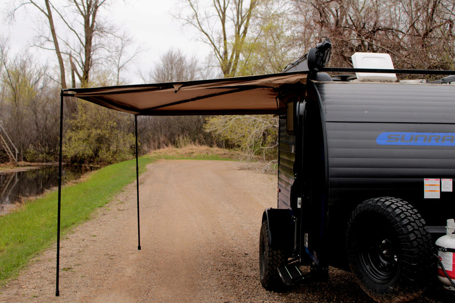 Black Sunray RV rental with extended awning, parked on a gravel road beside a pond. Ideal for your next outdoor adventure.