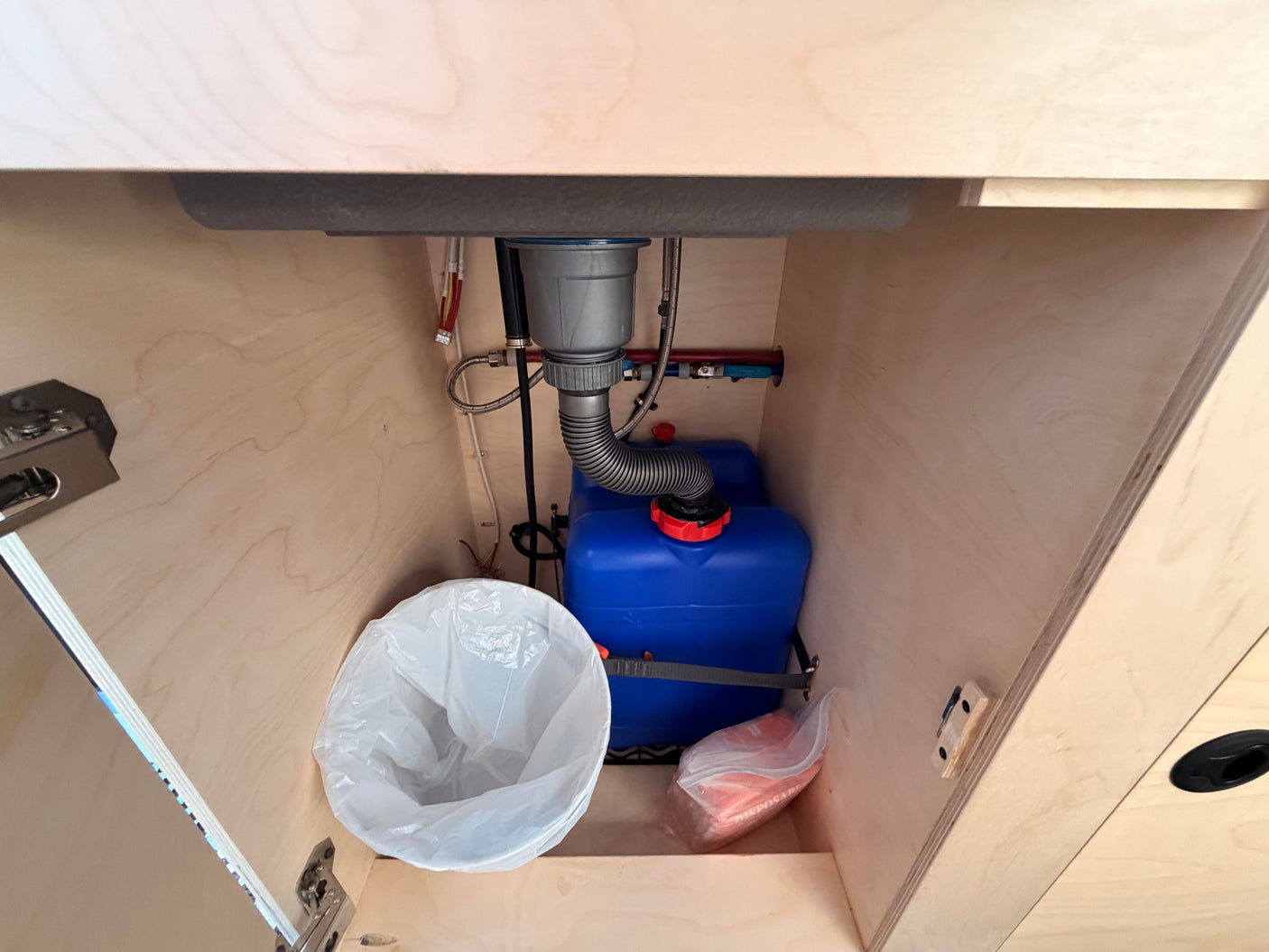 RV rental under-sink plumbing: view of a grey water tank, drain, and small trash bin inside a wooden cabinet.
