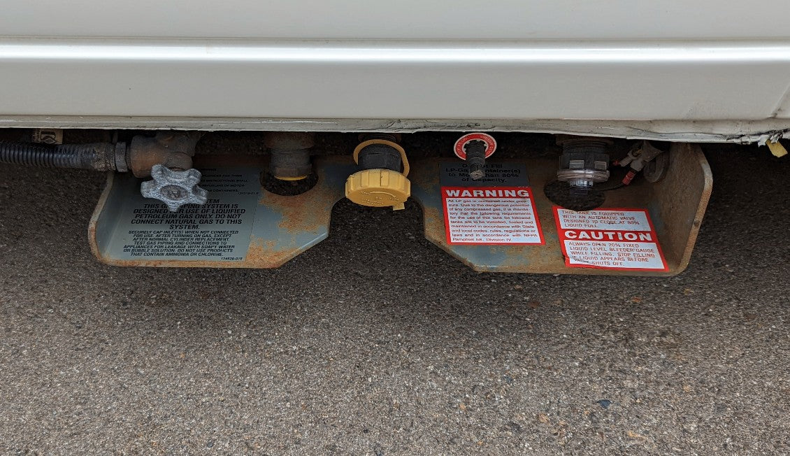 Underside view of an RV rental's LP gas system with propane tank, fill port, and safety labels for safe operation.