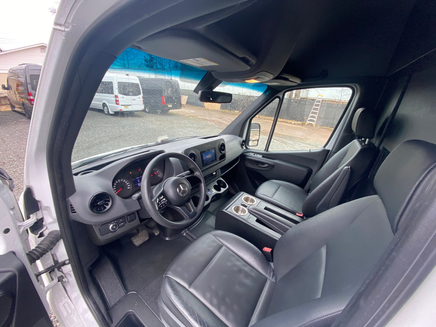 RV rental: Driver's seat view of a Sprinter van interior. Black leather seats, dashboard, and other vans outside are visible.