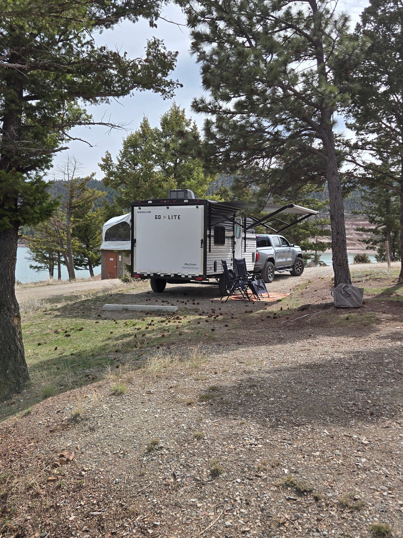 Lakeside rv rental setup with a white travel trailer, truck, and camping chairs under pine trees by the water.