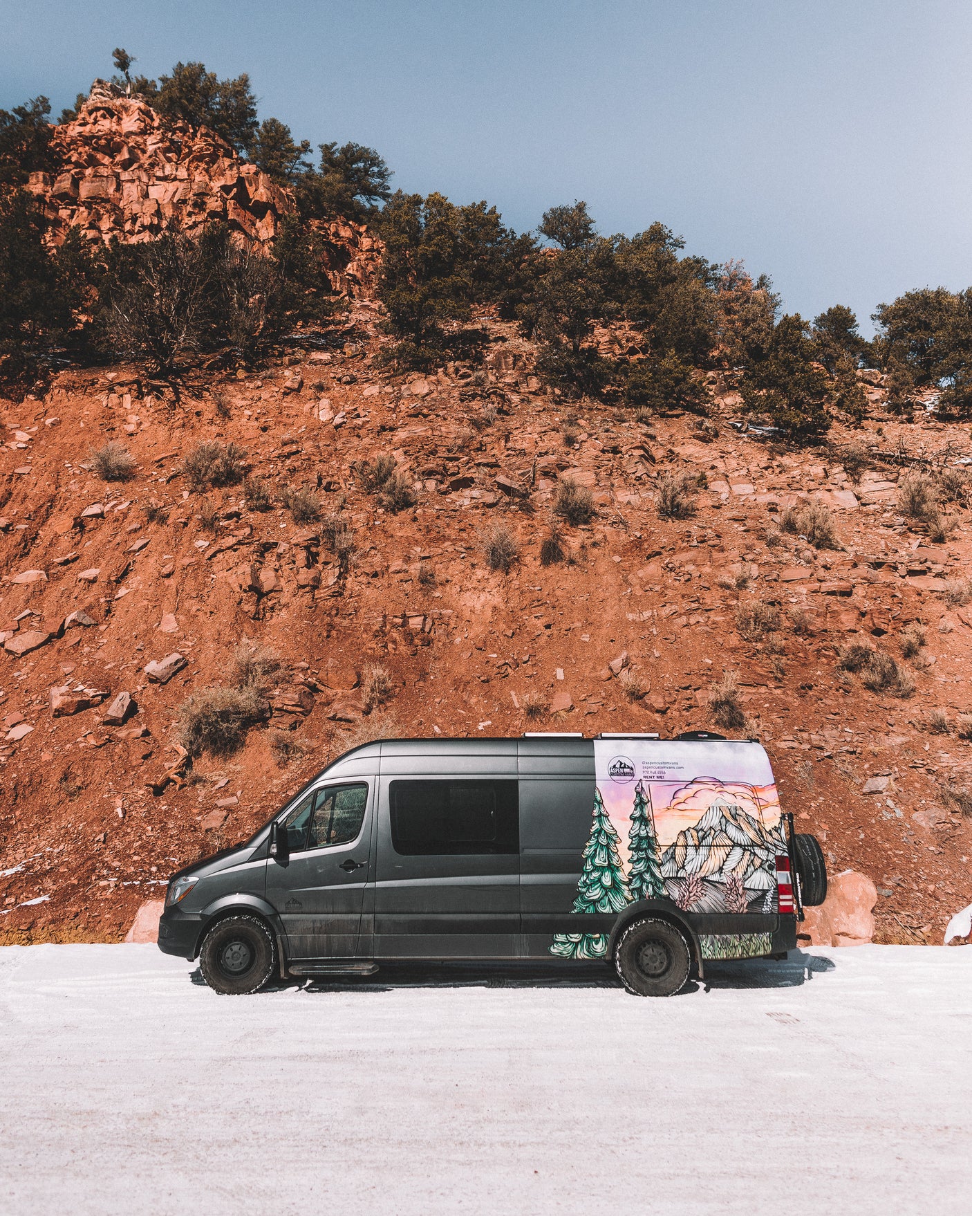 Dark grey RV rental van with custom mountain art, parked on a snowy road against a towering red rock cliff under a blue sky.