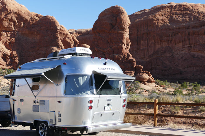Shiny Airstream rv rental parked in a sunlit desert. Its silver body contrasts with massive red rock formations and a clear blue sky.