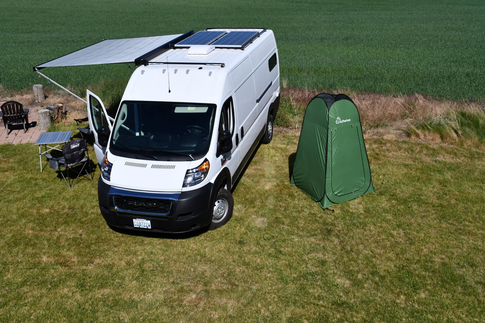 An rv rental white camper van with solar panels, extended awning, and a green privacy tent set up on a sunny grassy field.