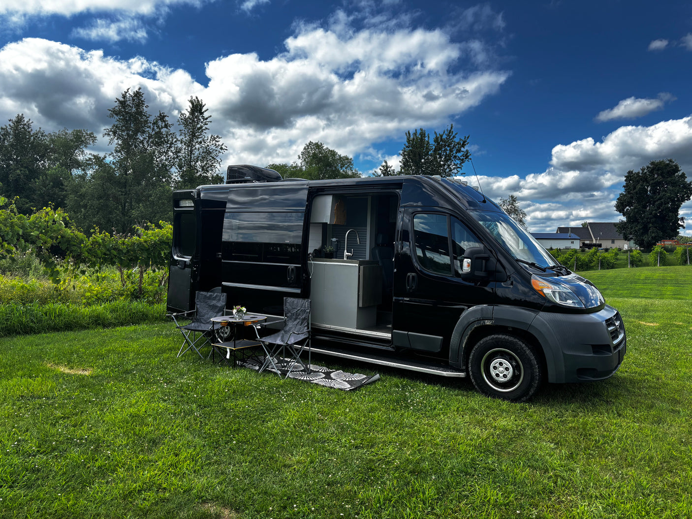 Black camper van RV rental in a vineyard, ready for outdoor living with chairs and table set up on lush grass.