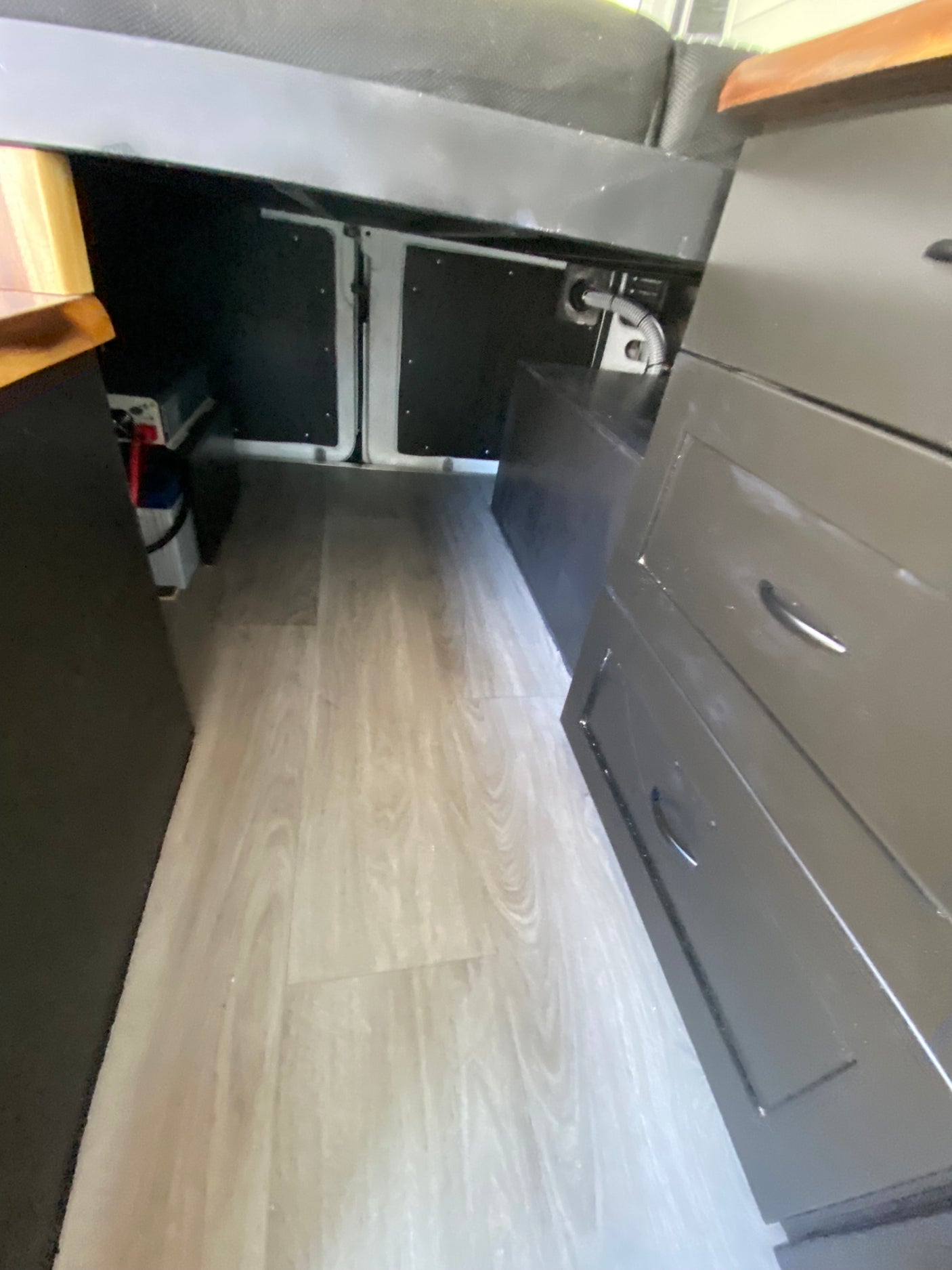 RV rental: Interior view showing light wood-look flooring, under-bed storage with batteries, and a dark cabinet with drawers.