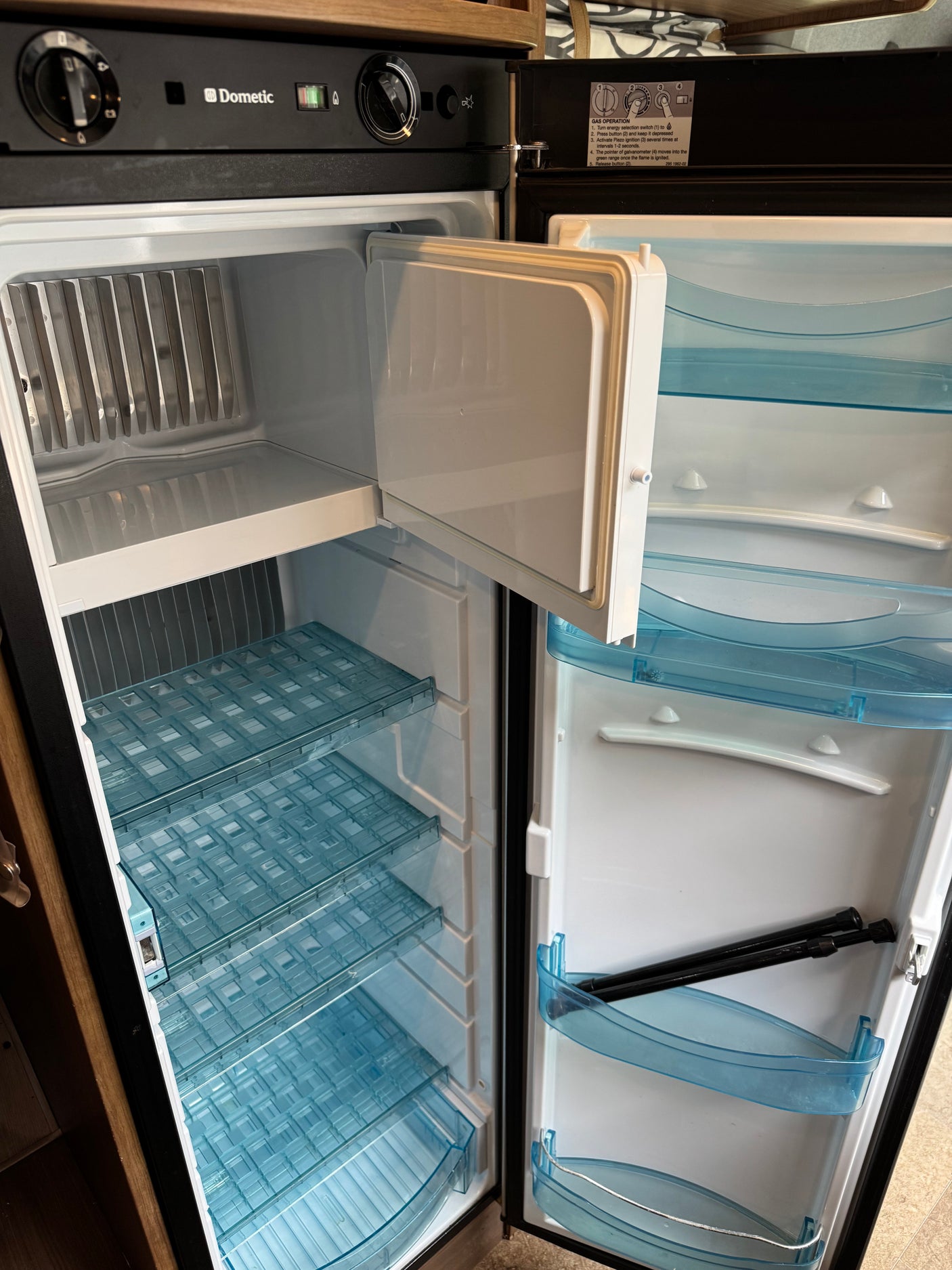 An empty Dometic RV refrigerator from an rv rental, showing the top freezer, blue gridded shelves, and door storage. Gas operation instructions are visible.