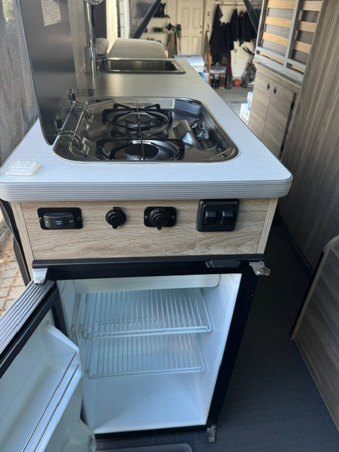 An rv rental's compact kitchen features a two-burner stove, sink with faucet, and an open mini-fridge with shelves.