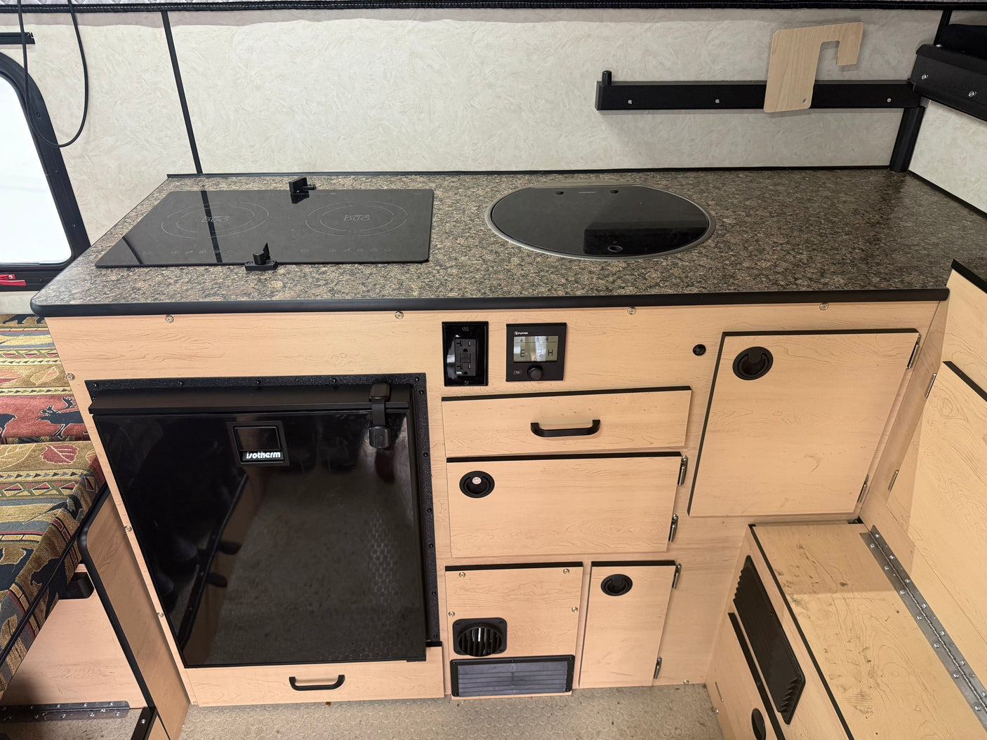 Modern rv rental galley kitchen with granite-look countertop, induction cooktop, covered sink, Isotherm fridge, and Truma controls.