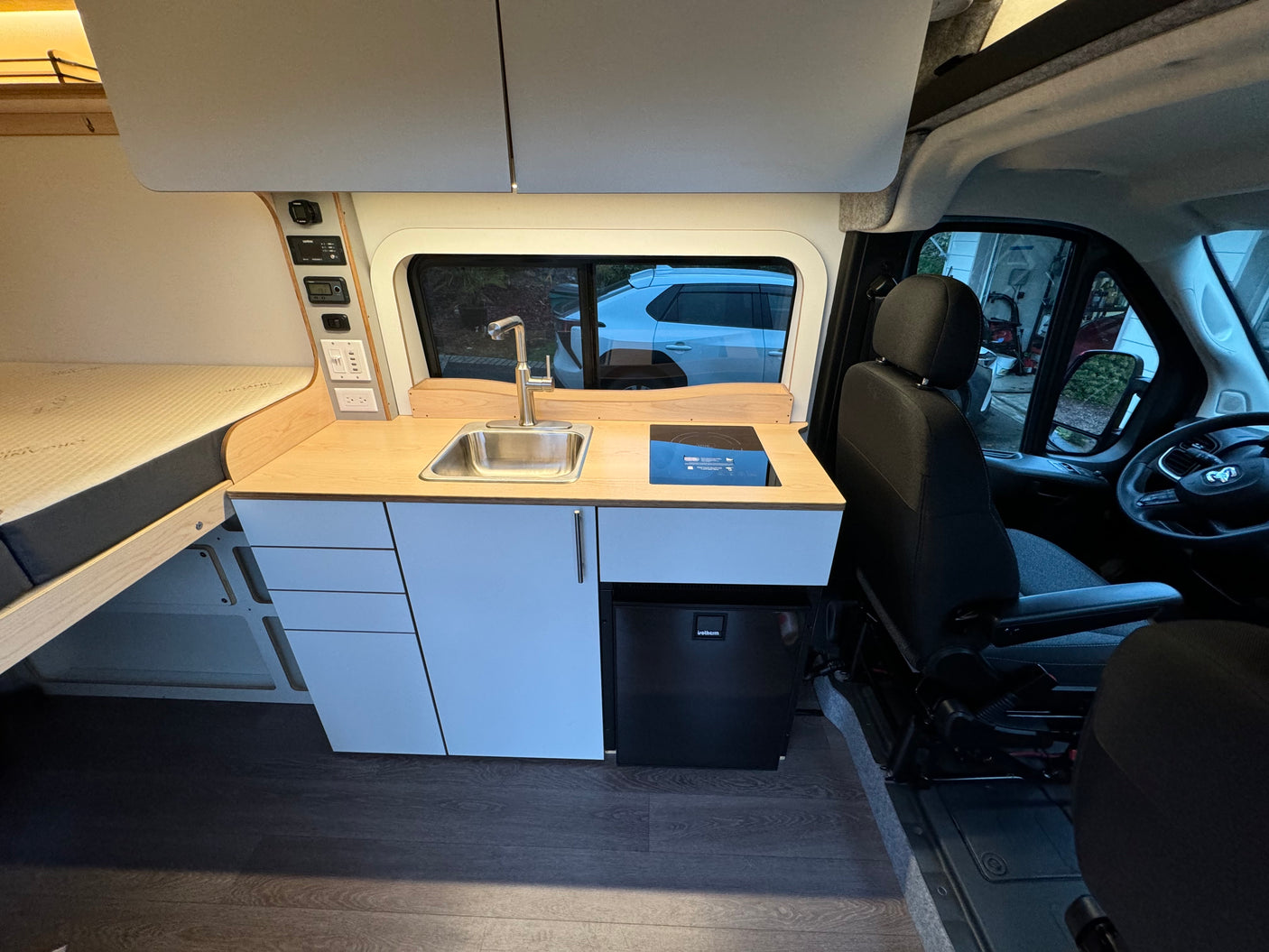 This modern RV rental interior showcases a fully equipped kitchenette with a sink, cooktop, and fridge, plus a comfortable bed and control panel.