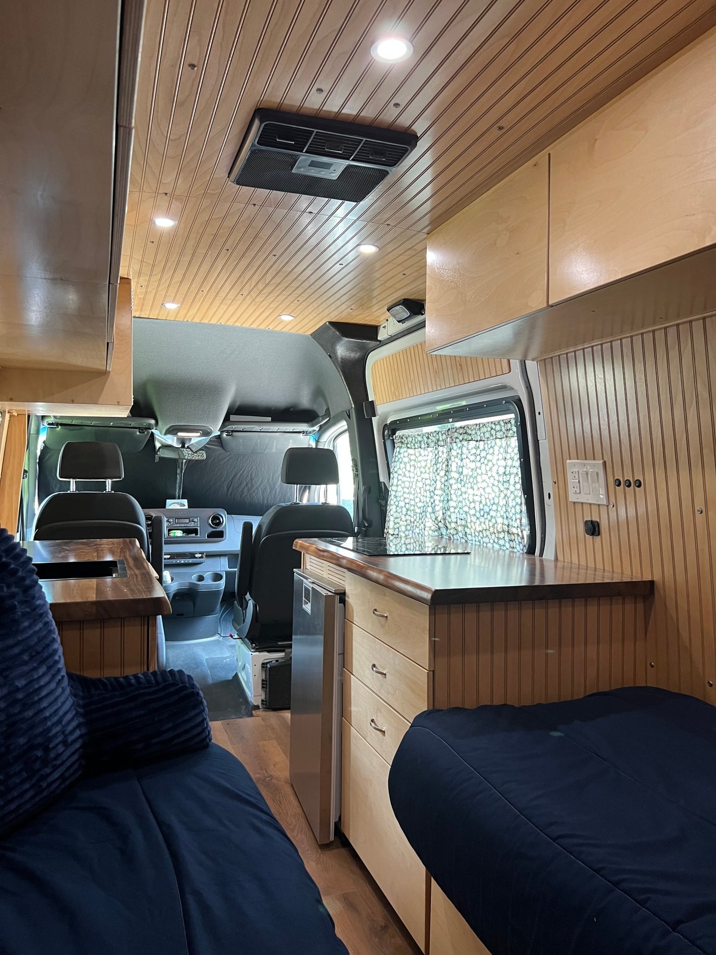 Spacious custom RV rental interior with wood paneling, recessed lighting, kitchen area, and comfortable seating, extending to the driver's cab.