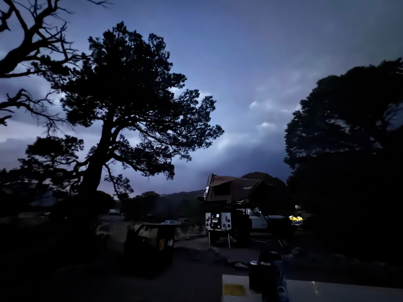 An RV rental with a rooftop tent parked at a dark campsite under a cloudy night sky, surrounded by large silhouetted trees.