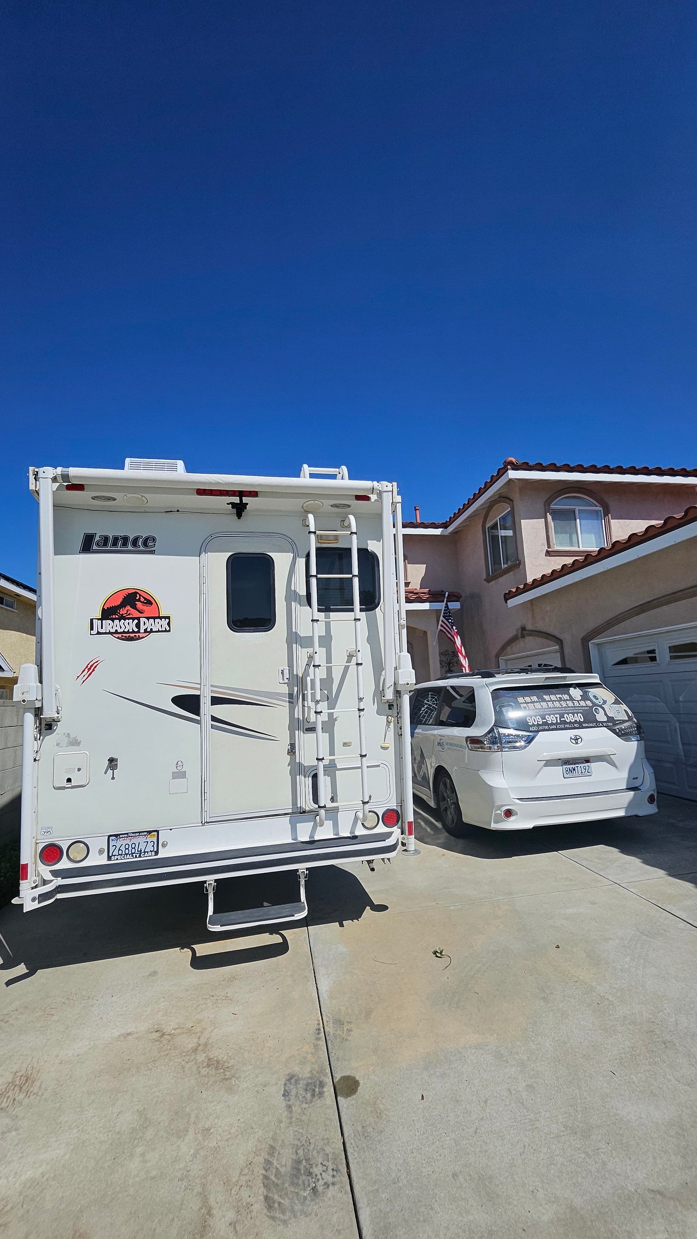 A white rv rental with a Jurassic Park decal is parked in a driveway next to a minivan and a house on a clear day.