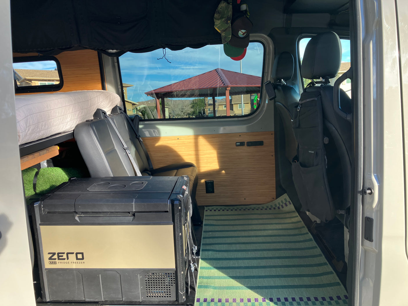RV rental: View inside a camper van with an ARB Zero fridge freezer, bed, passenger seats, and custom wood panels.