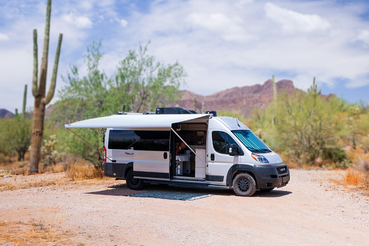 A white camper van RV rental with an extended awning is parked on a dirt road in a sunny desert landscape