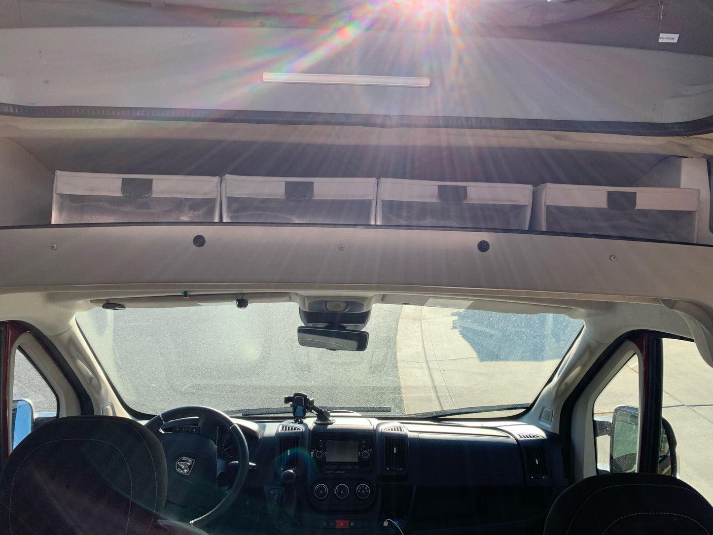 View from inside an RV rental: driver's area, dashboard, windshield, and overhead storage bins. Sunlight flares.