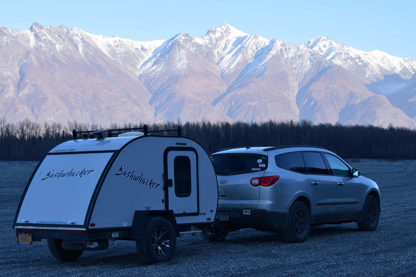 Bushwhacker RV rental and SUV on gravel, against majestic snow-capped mountains and bare trees.
