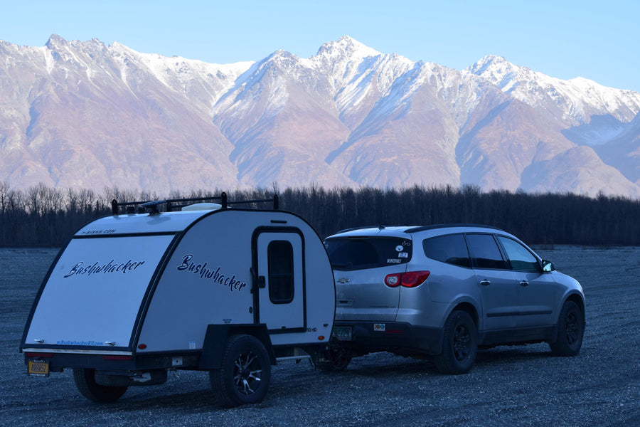 Bushwhacker RV rental and SUV on gravel, against majestic snow-capped mountains and bare trees.