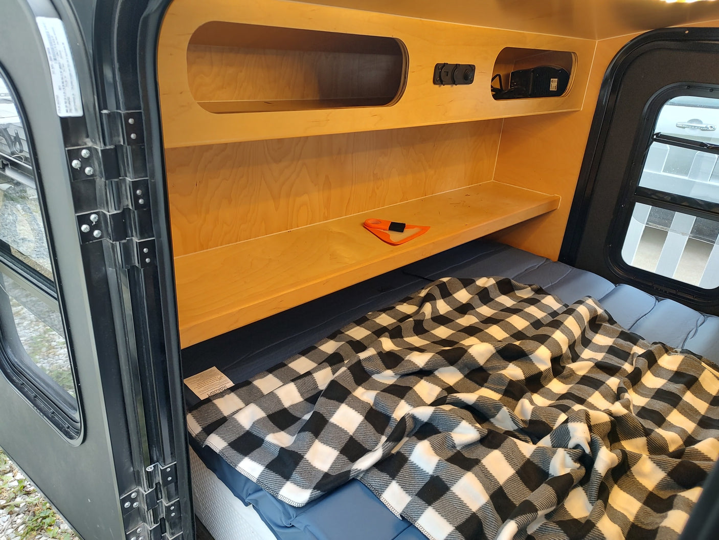 Explore this RV rental's comfy interior: a bed with checkered blanket, built-in wooden storage, and a large window.