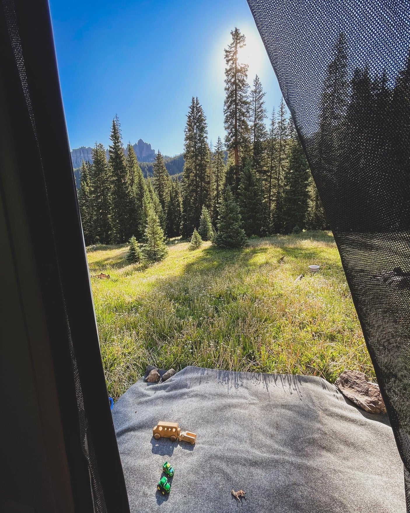 Morning view from an RV rental, looking out at a sunny forest meadow with mountains. Kids' toys on the blanket inside. Perfect camping!