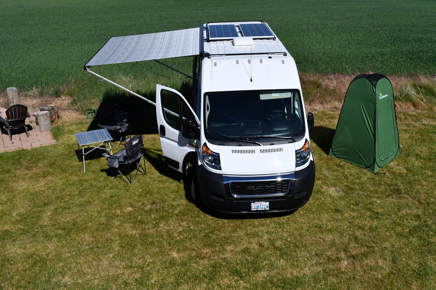 White **rv rental** van with an extended awning & solar panels, surrounded by camping chairs, table, & a green privacy tent on grass.