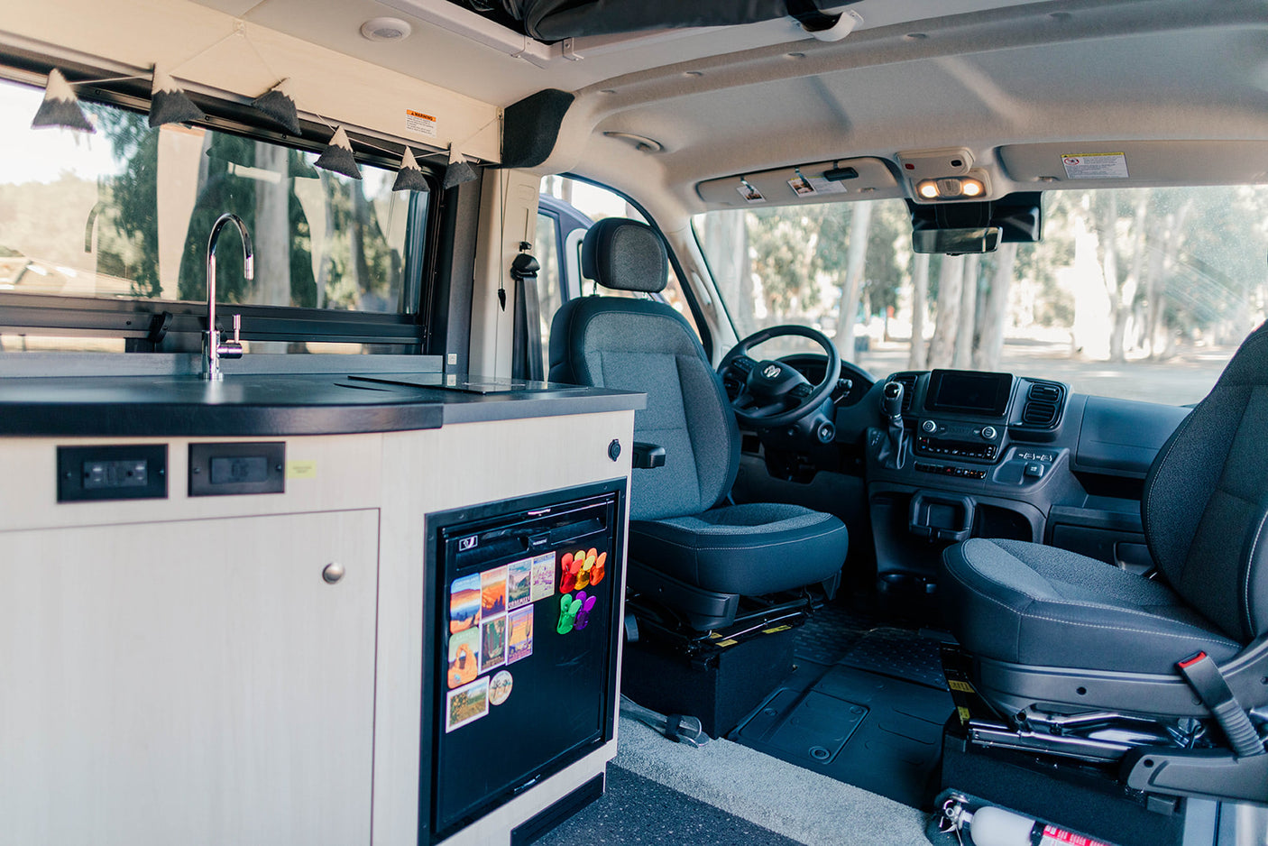RV rental interior: light wood kitchenette with sink, fridge, and decorative mountain garland. Driver's cabin in view.
