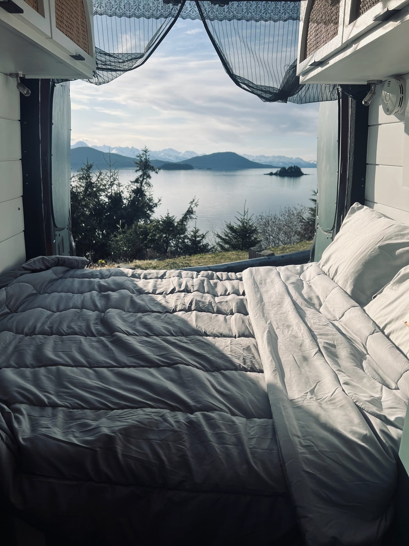 An RV rental bedroom with a view! Cozy bed overlooks a serene lake, evergreen trees, and snowy mountains. Perfect travel.