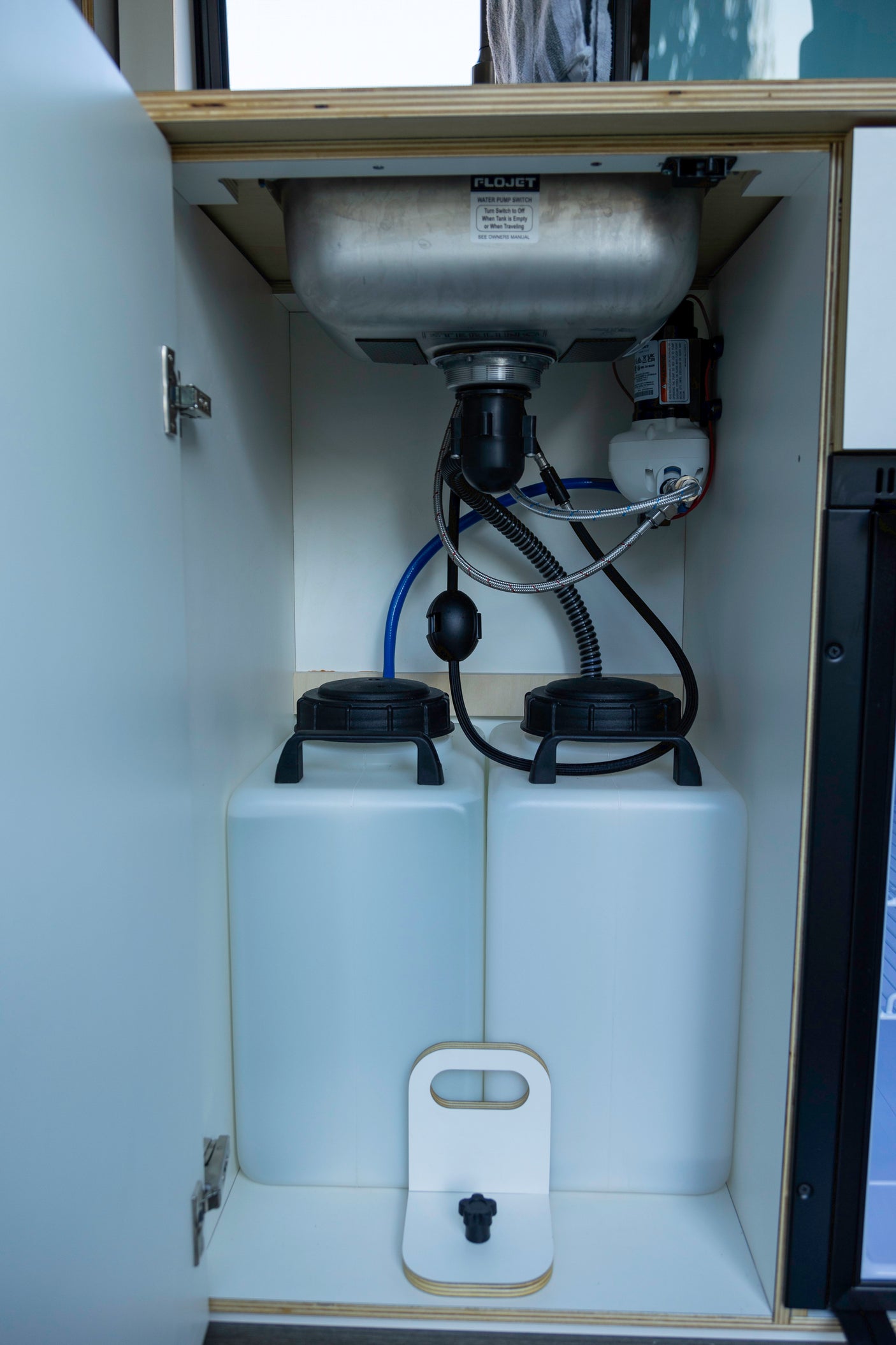 RV rental under-sink cabinet showing compact water system with two tanks, pump, and plumbing for a mobile kitchen.