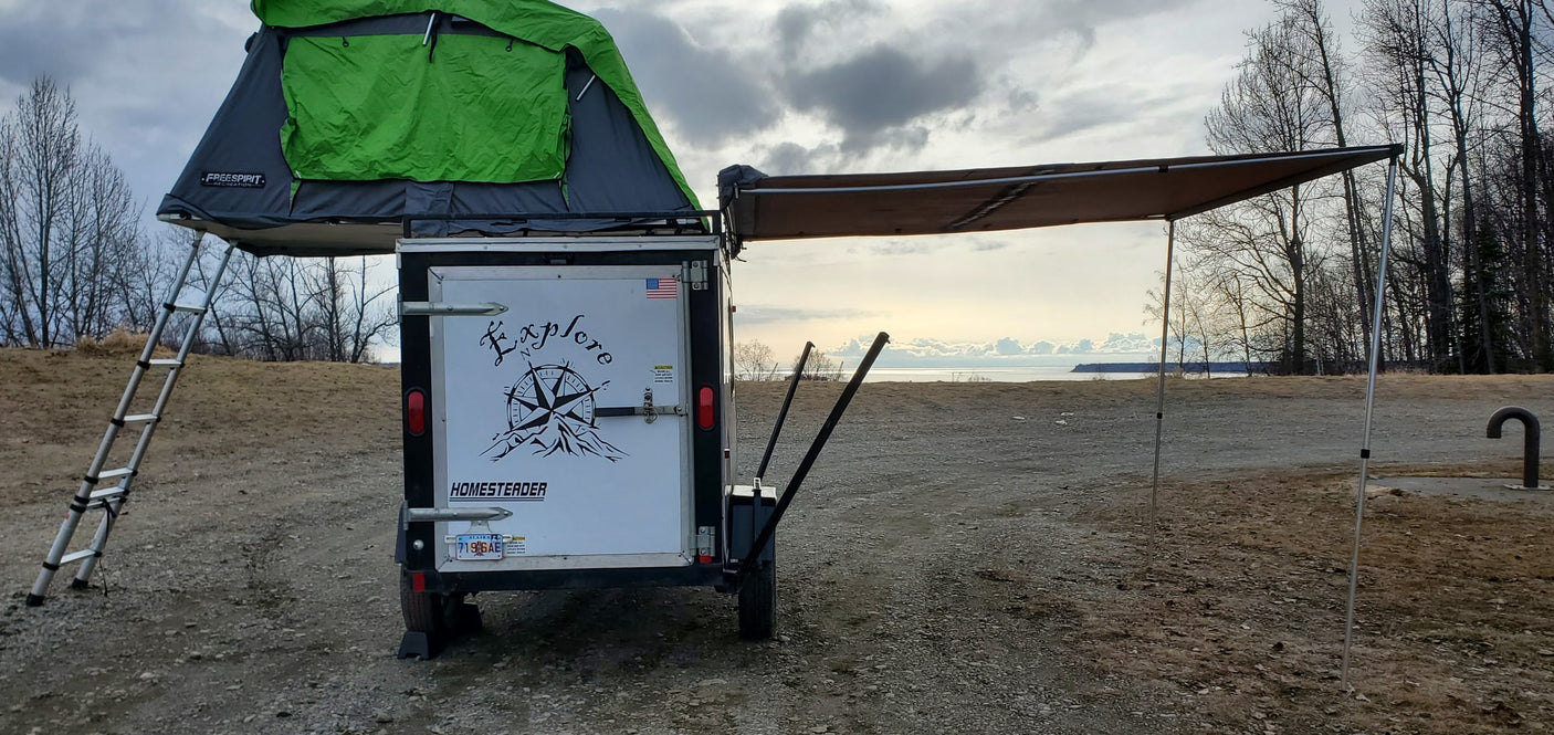 RV rental-style setup: a small trailer with a rooftop tent, ladder, and awning by a large body of water under a cloudy sky.
