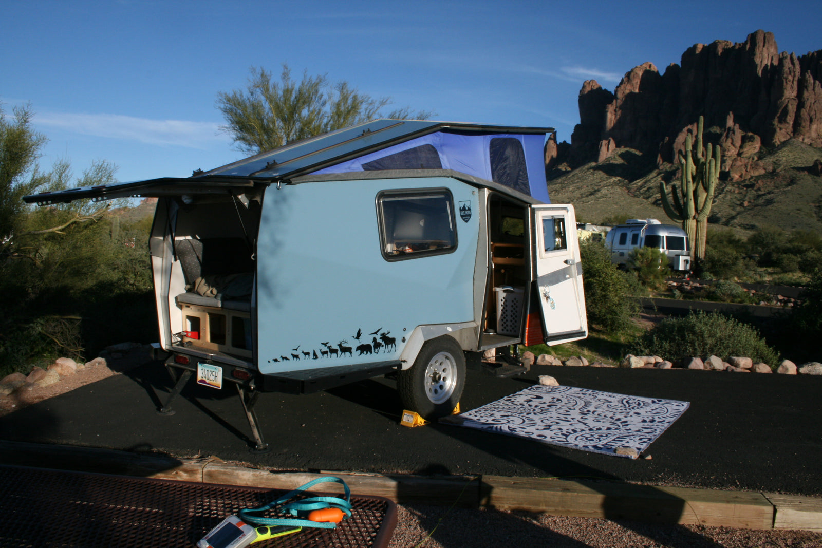 Enjoy desert adventures! This compact teardrop RV rental, with its pop-top tent, offers cozy camping amidst mountains & cacti.