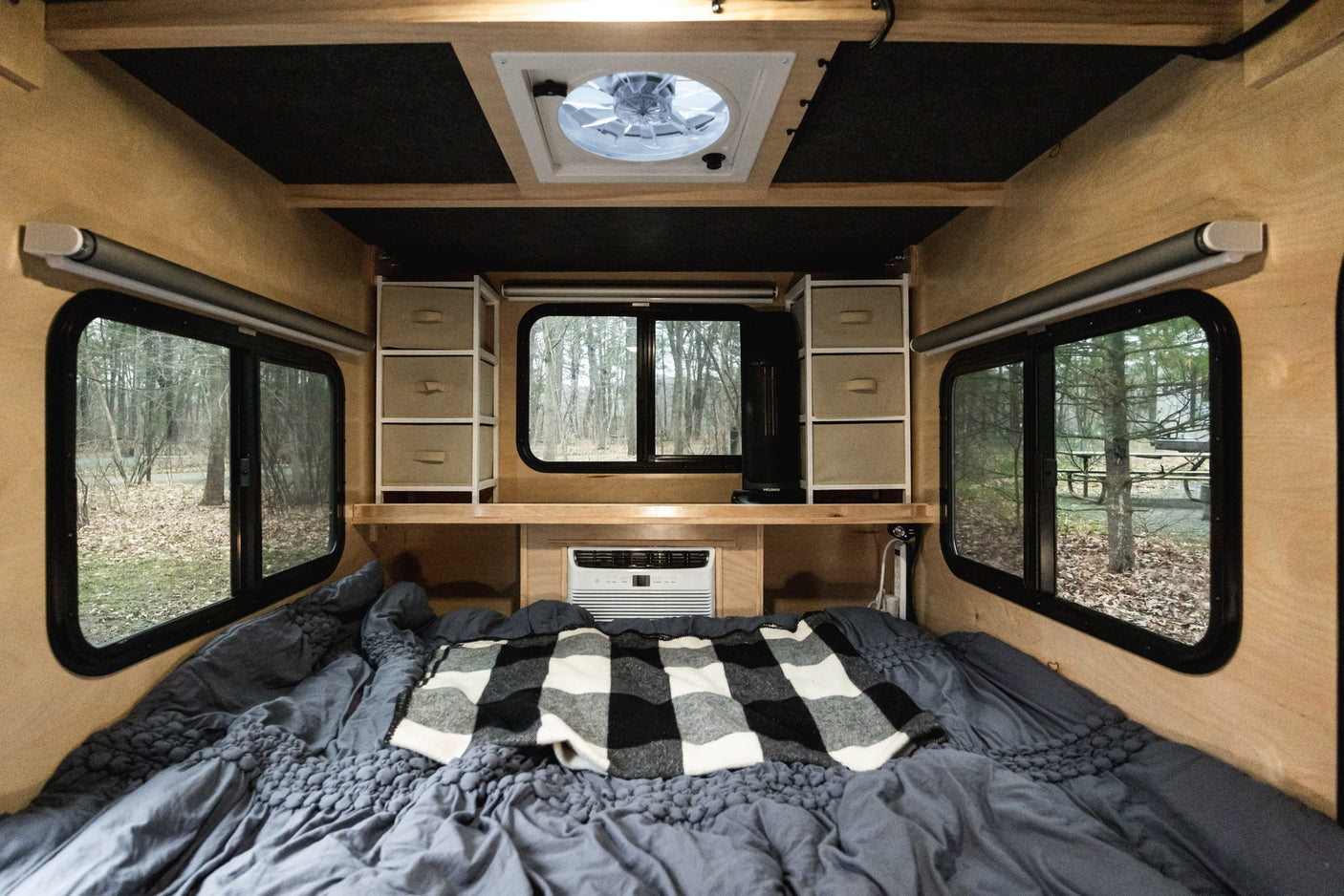 Cozy RV rental interior with comfortable bed, overhead storage, AC, and serene forest views through large windows.