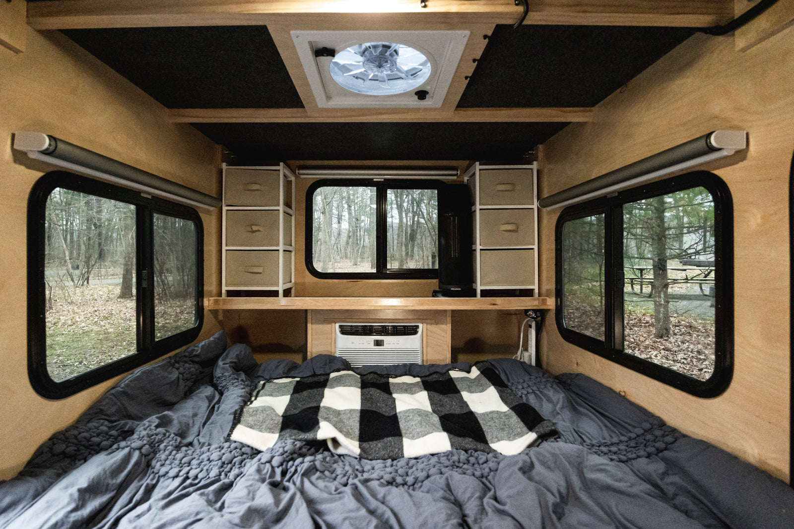 Cozy RV rental interior with comfortable bed, overhead storage, AC, and serene forest views through large windows.
