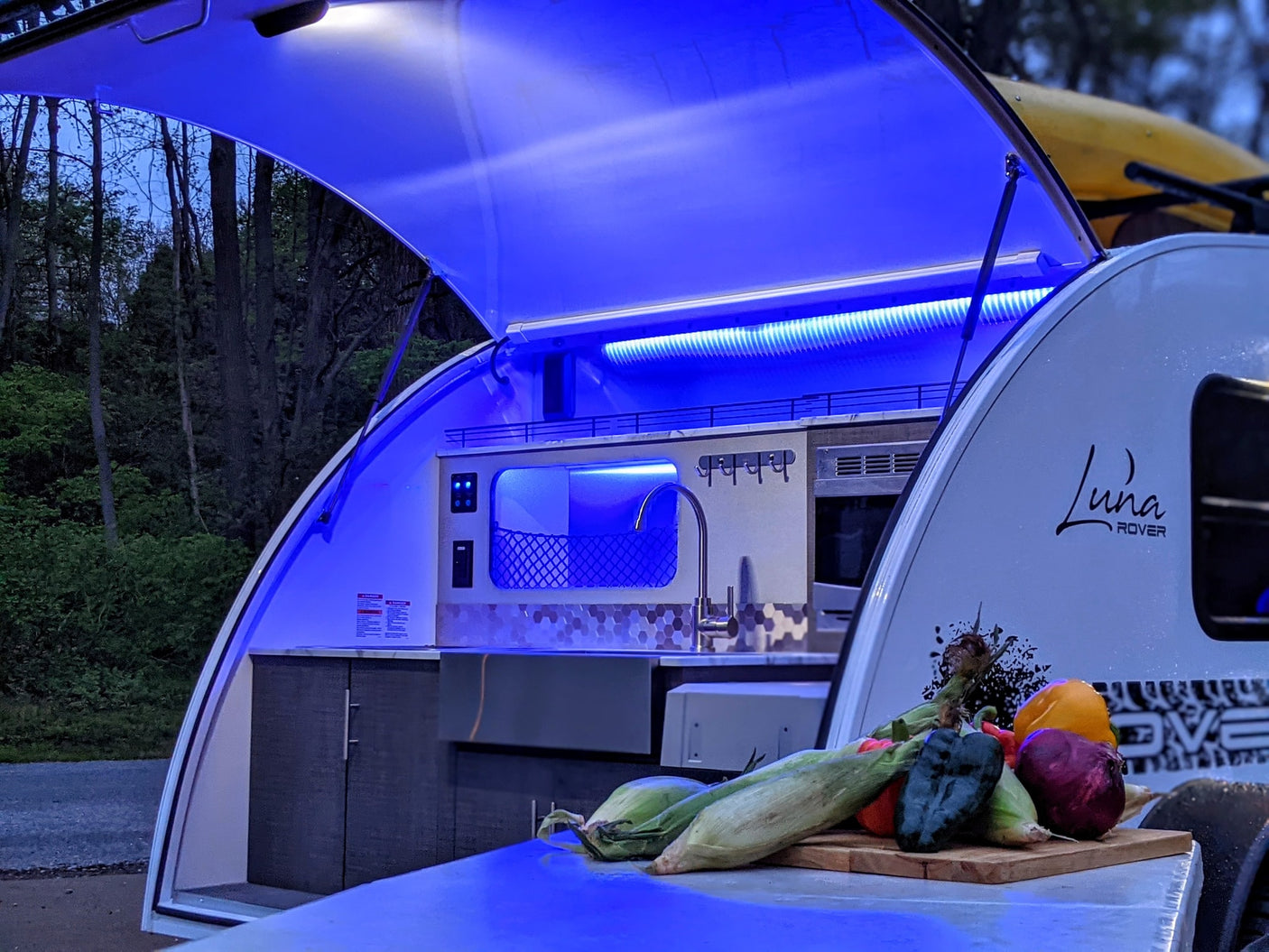 Teardrop camper RV rental with illuminated outdoor kitchen for camping cooking. Fresh veggies ready for a meal.