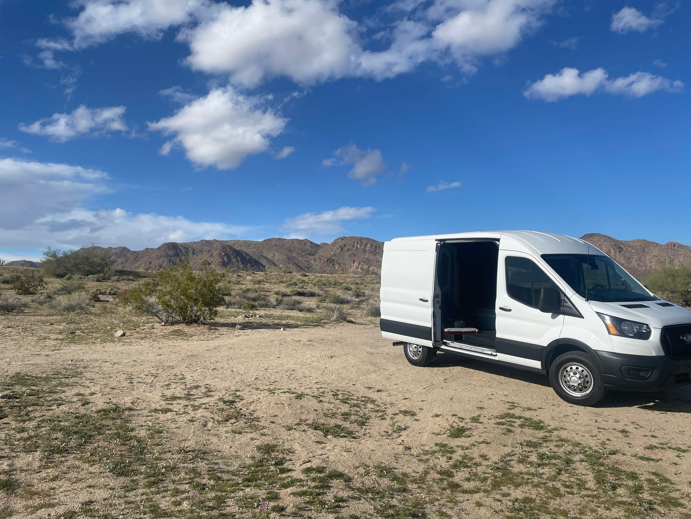 White Ford Transit RV rental in a vast desert landscape with mountains and blue sky, side door open.