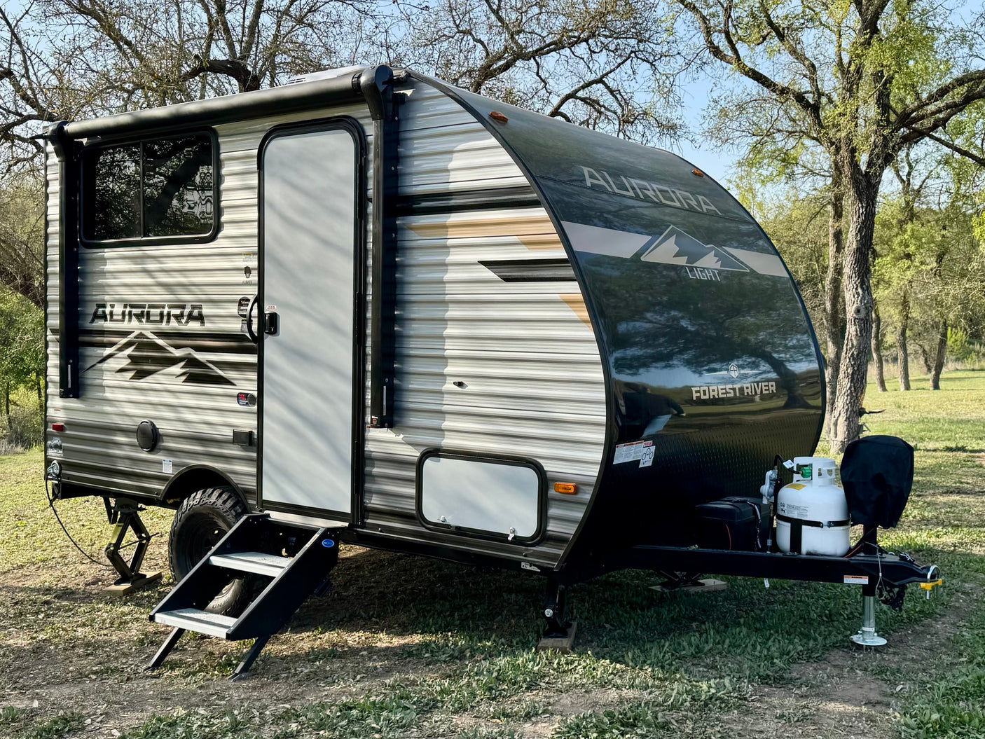 A sleek Forest River Aurora travel trailer, ideal for your next rv rental, parked on grass amidst mature trees.