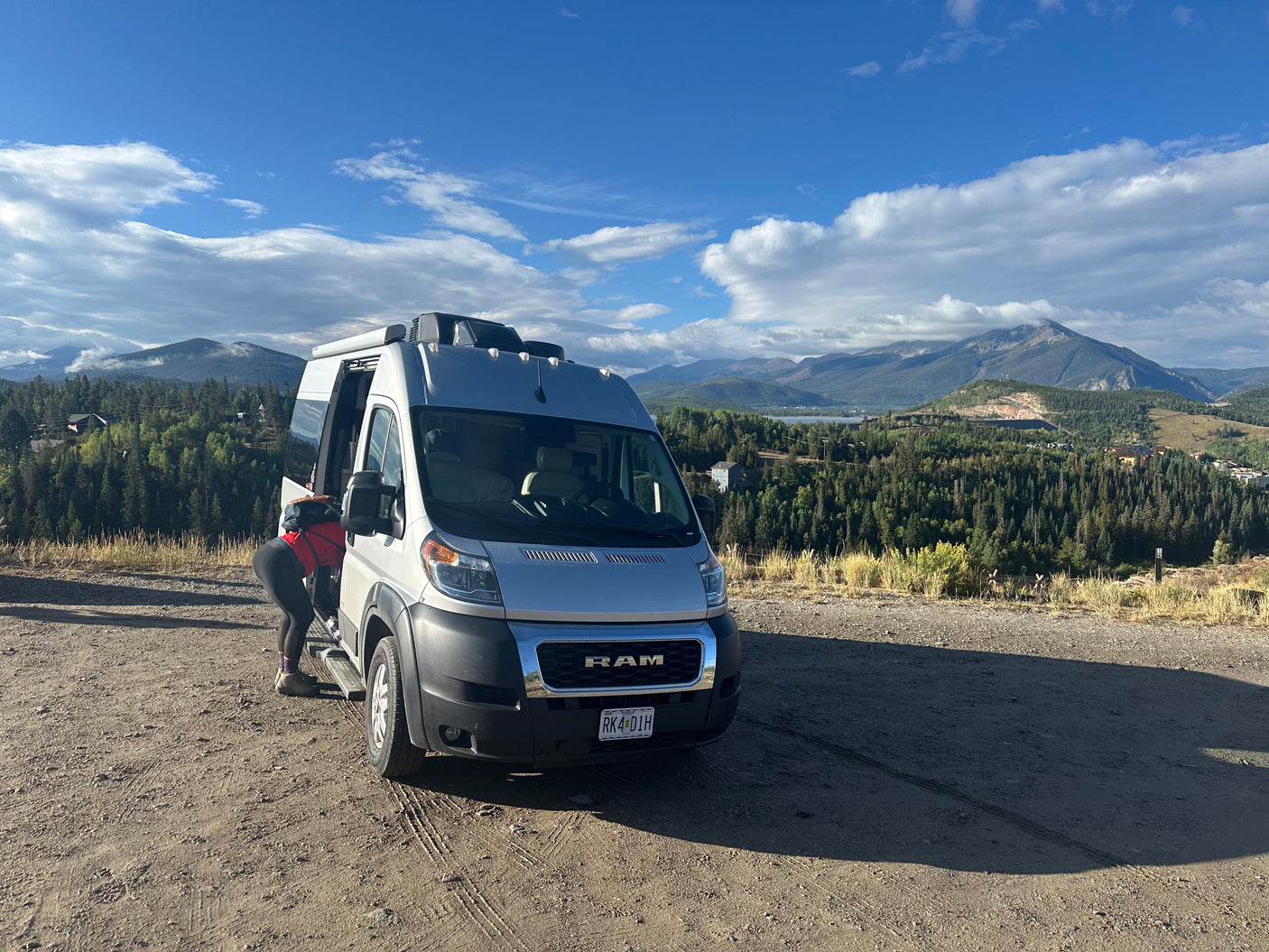 An RV rental camper van stopped on a mountain overlook. A person accesses the van, revealing a breathtaking view of forested peaks and blue sky.