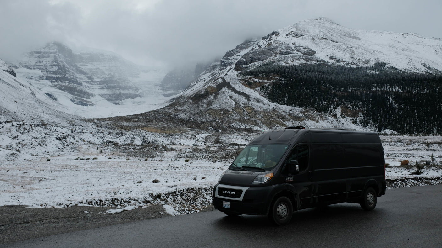 Scenic winter landscape with a black RV rental van parked on a wet road amidst majestic, snow-covered mountains.