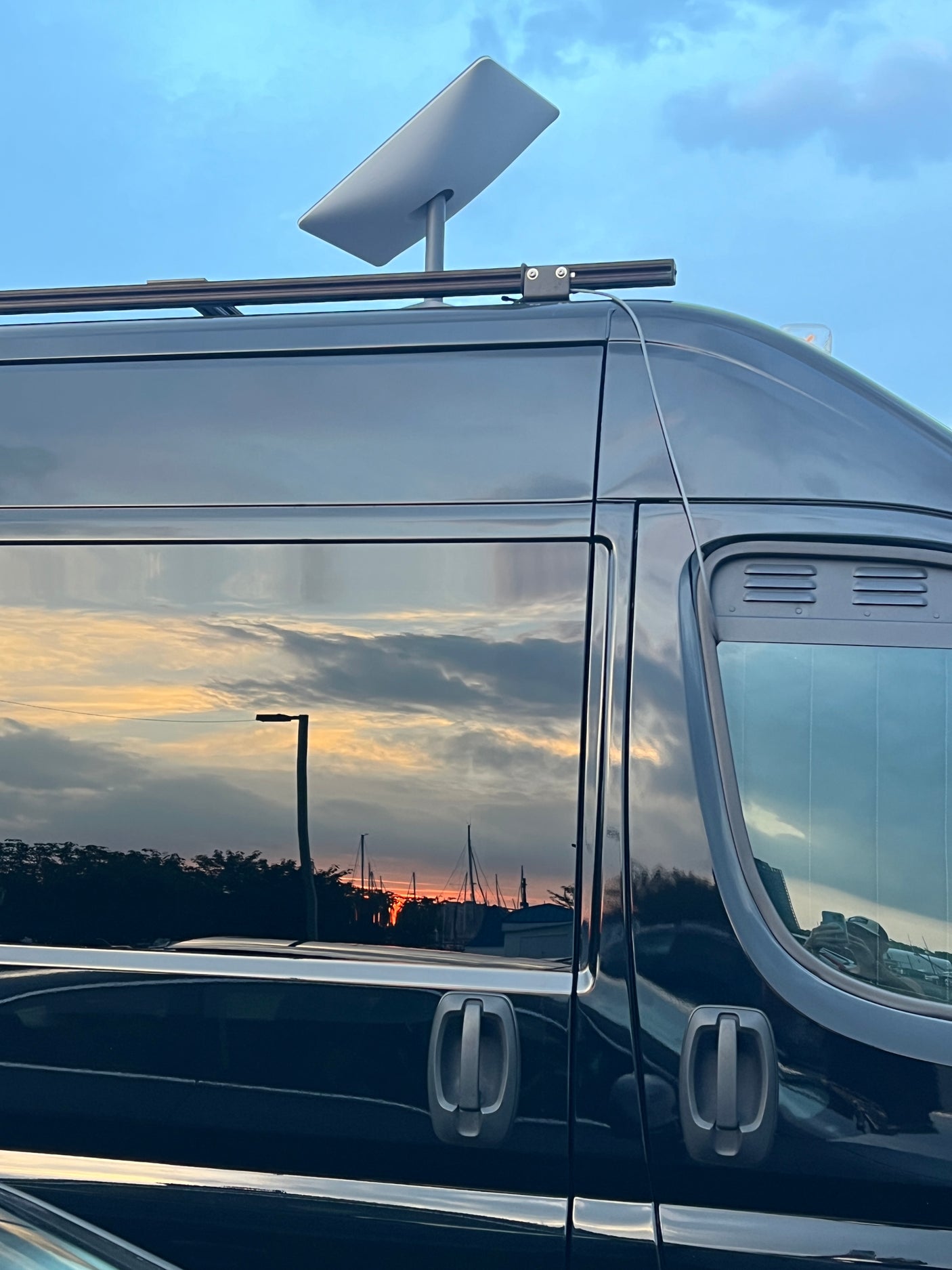 Black RV rental with a Starlink satellite dish mounted on its roof rack. Sunset with boats reflected in the side windows.