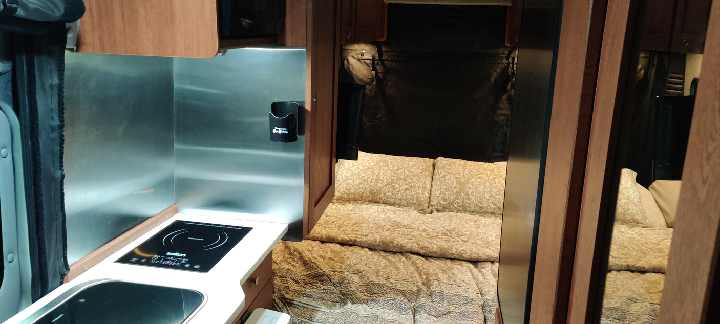 This RV rental interior features a compact kitchen with induction cooktop, sink, and a cozy bedroom with patterned bedding.