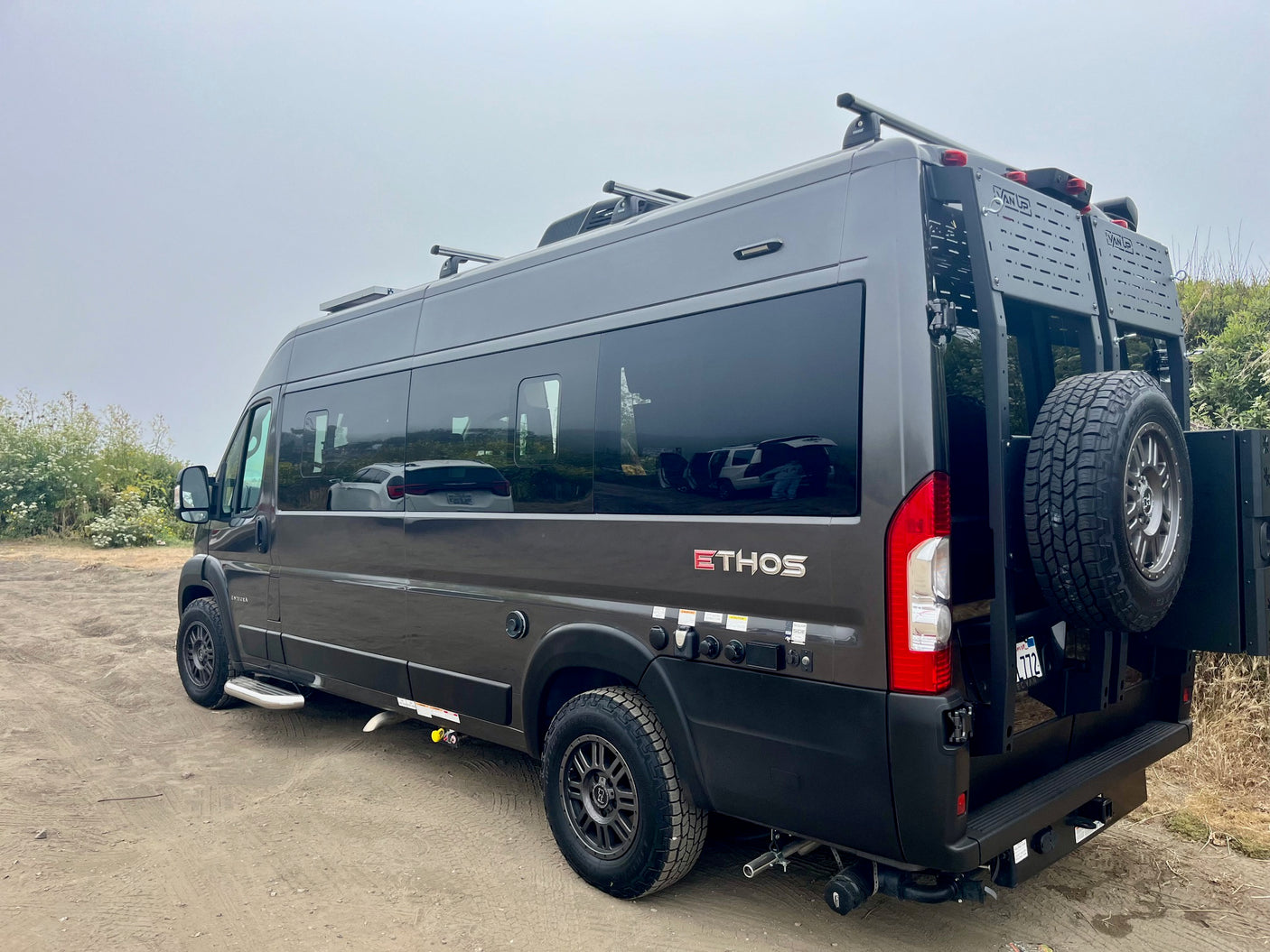 Dark grey Ethos overland camper van, equipped for off-road rv rental. Features roof rack & rear gear carrier.