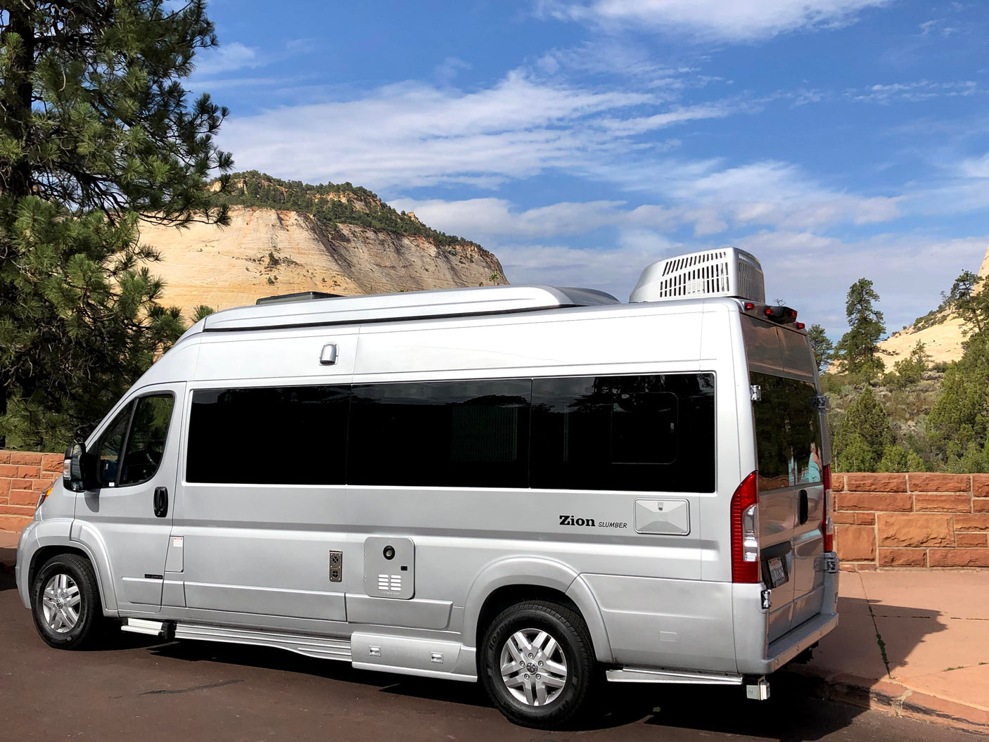 Silver camper van RV rental, a 'Zion Slumber' model, parked against a scenic backdrop of layered mountains and blue sky.